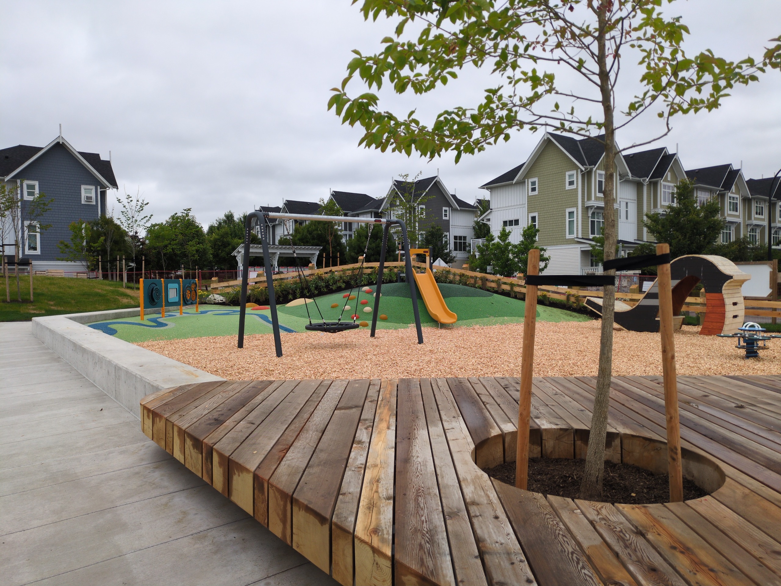 A heavy timber wood deck built around a tree in a central plaza, with playground behind.