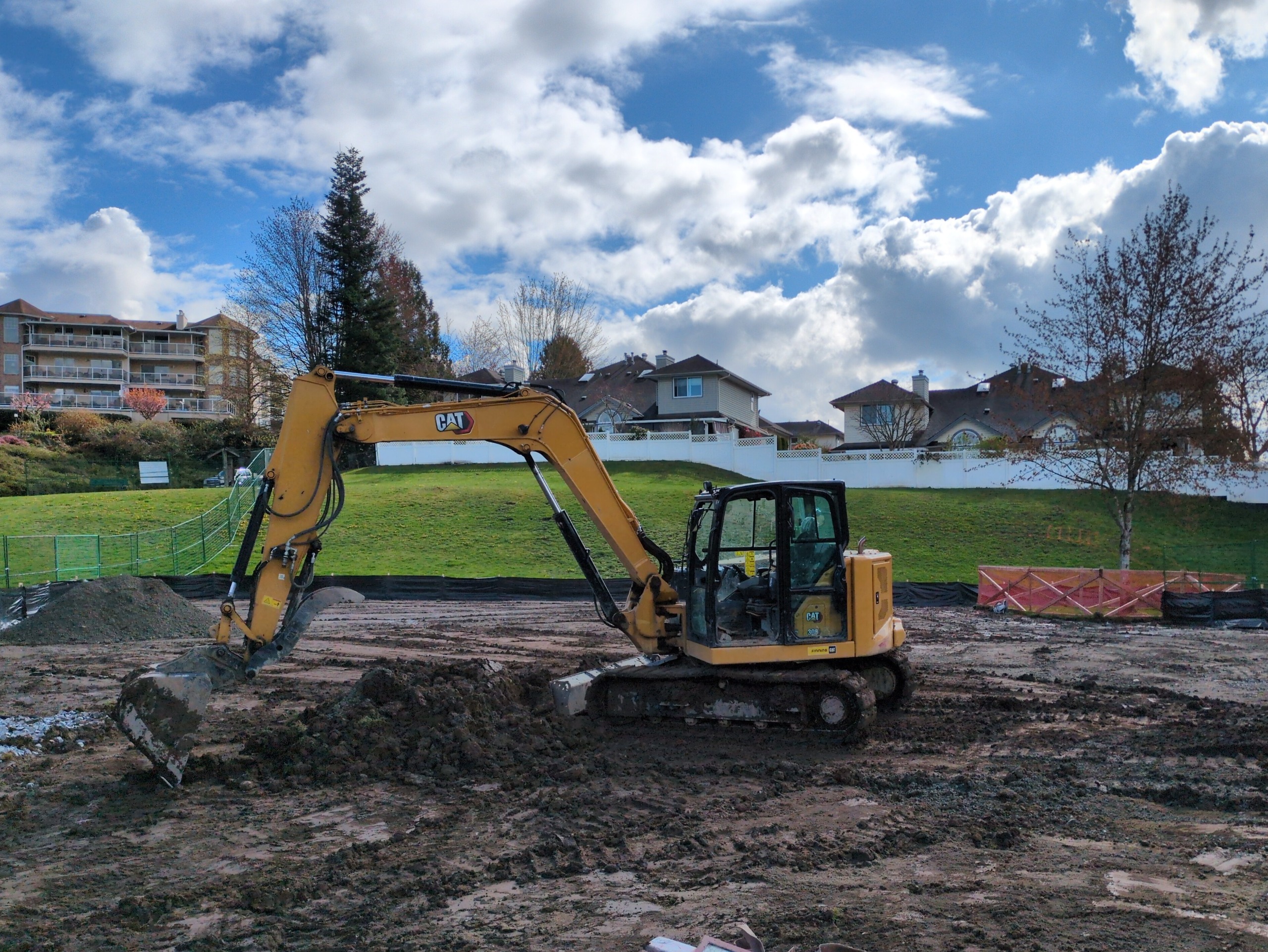 An excavator in a construction site with sod removed and tree protection fencing visible in the background