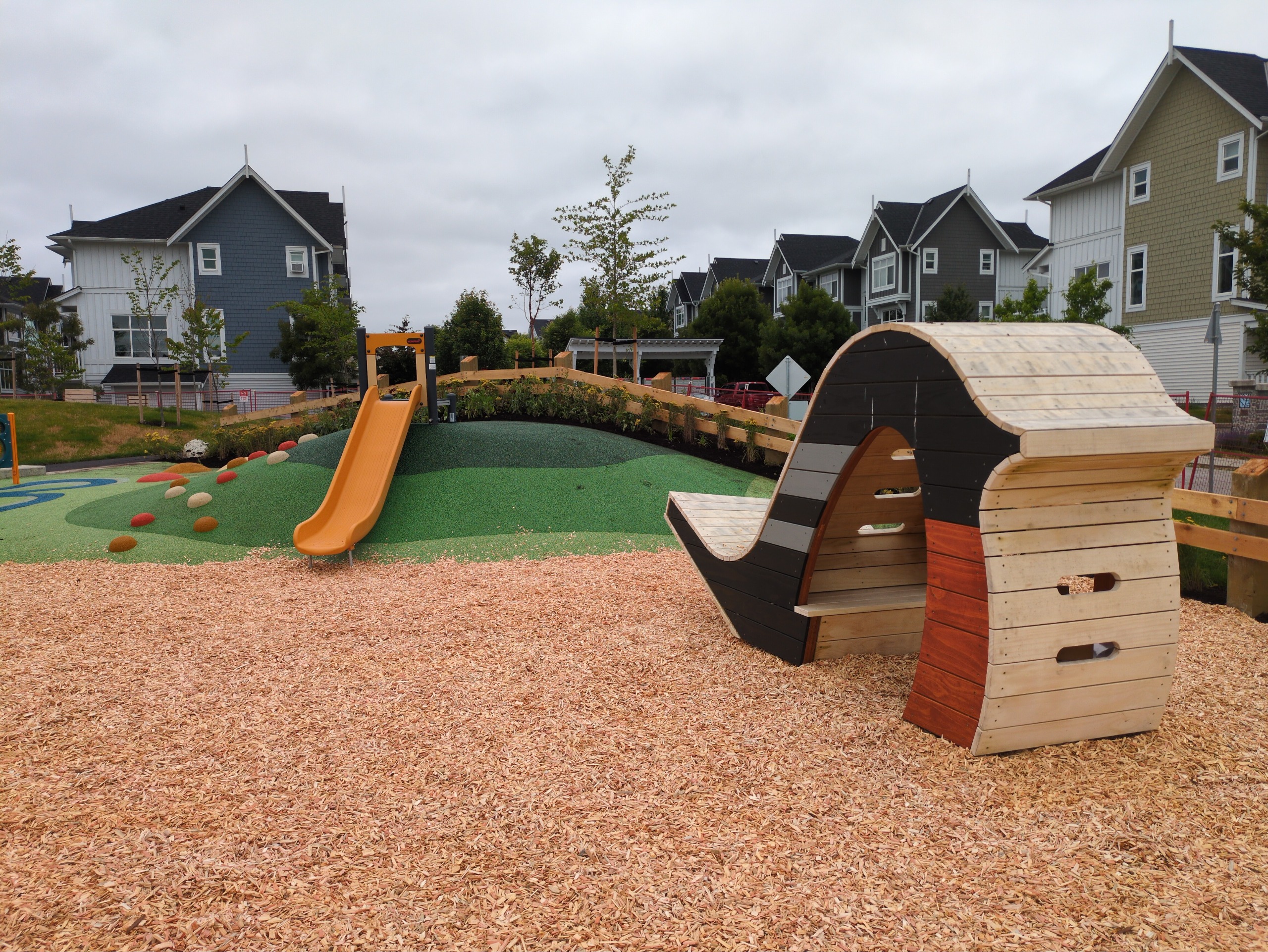 A wooden bird-shaped play hut on the right with a yellow hill slide on a green rubber hill on the left